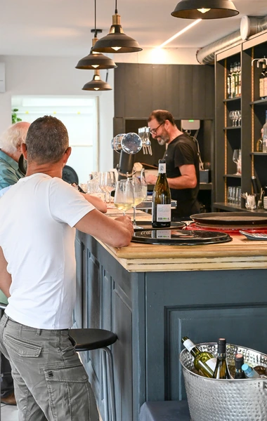 Clients au comptoir du bar des Platanes, échangeant avec le barman dans une ambiance conviviale.